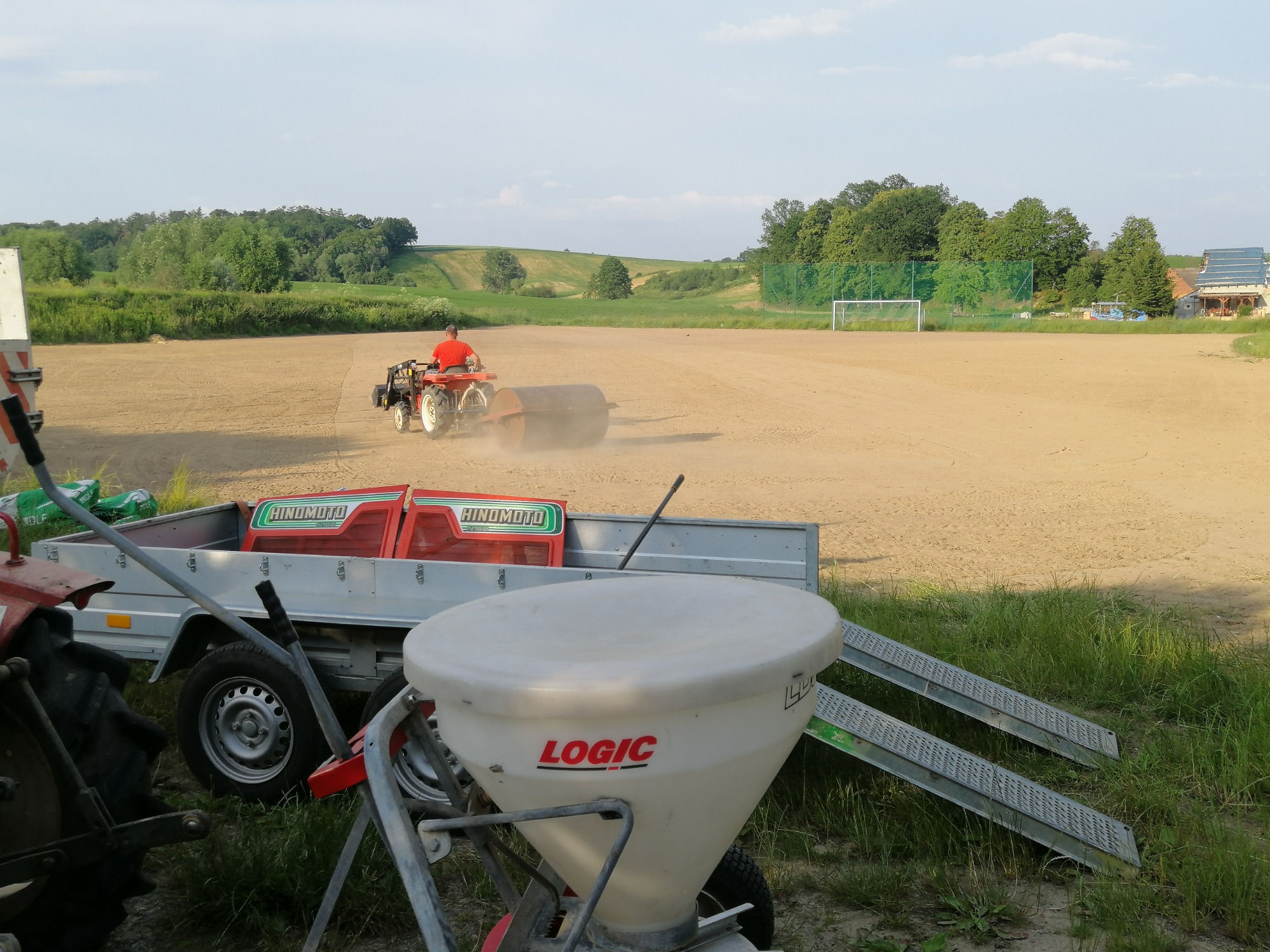 Operator na małym ciągniku z wałem wyrównuje powierzchnię piaszczystego boiska sportowego; w tle widoczna siatka bramki i zadrzewione wzgórza.