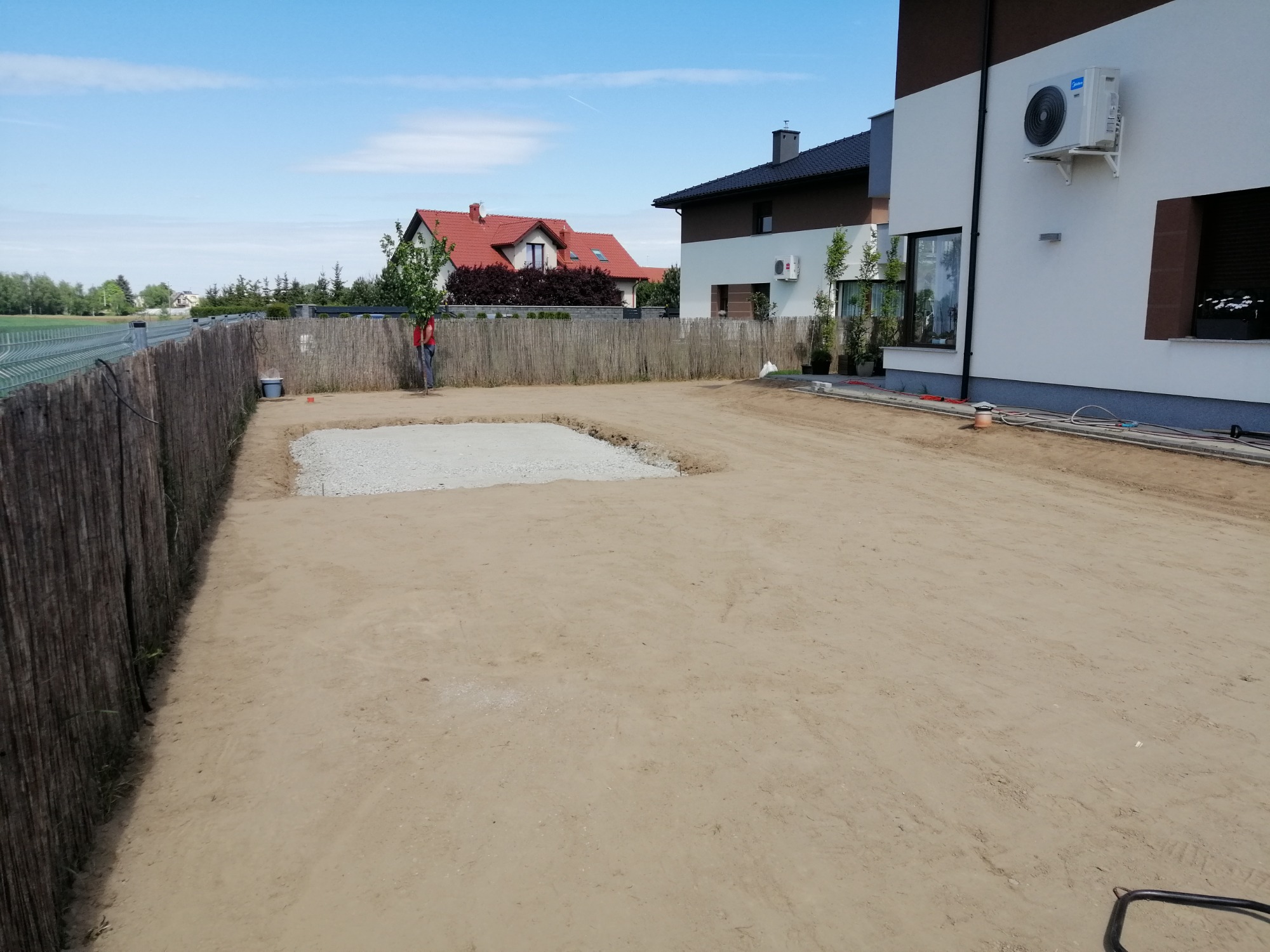 Przygotowany teren pod budowę z wykopanym fundamentem, widoczny pracownik przy ogrodzeniu i domy w tle.