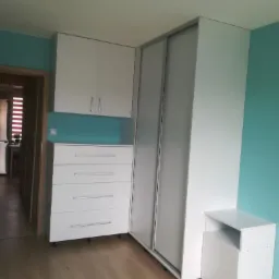 Biała szafa narożna z przesuwnymi drzwiami, komodą z szufladami i szafką nocną na kółkach, dopasowana do turkusowej ściany.