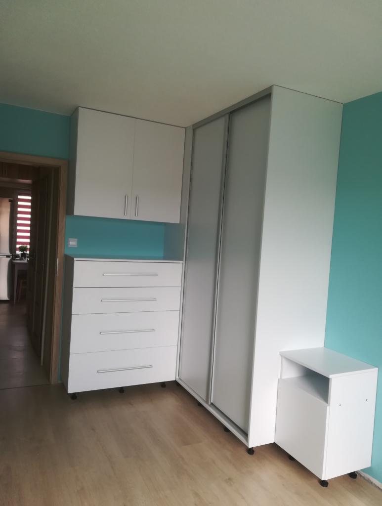 Biała szafa narożna z przesuwnymi drzwiami, komodą z szufladami i szafką nocną na kółkach, dopasowana do turkusowej ściany.