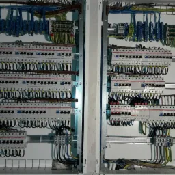 Dwie rozdzielnice elektryczne z widocznymi bezpiecznikami, przewodami i listwami zaciskowymi, zamontowane w białych obudowach, z przewodami ułożonymi w regularne pętle.