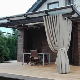 Zadaszony taras z drewnianą podłogą, zasłonięty beżowymi, grubymi zasłonami spiętymi sznurami, z widocznym stołem i krzesłami w tle, oświetlony dekoracyjnymi żarówkami.