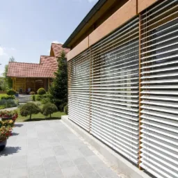 Elewacja domu z drewnianymi detalami i zamontowanymi białymi poziomymi osłonami okiennymi, widok z zewnątrz na tle zadbanego ogrodu z kostką brukową i roślinnością.