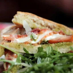 Kanapka z ciabatty z pesto, mozzarellą, pomidorem, szynką parmeńską i rukolą, posypana parmezanem, serwowana na drewnianej desce.