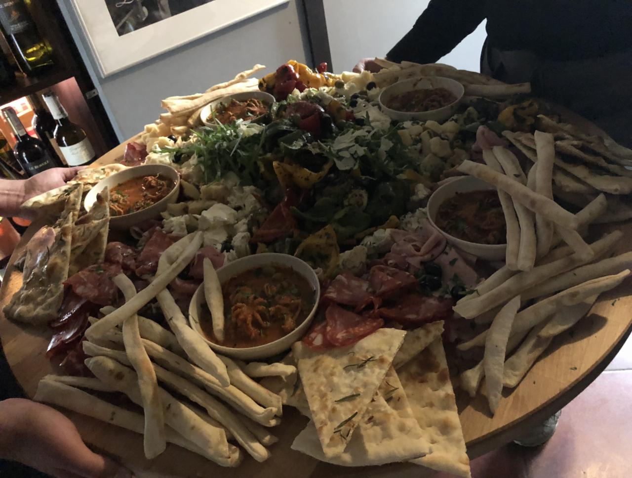 Duży drewniany talerz z różnorodnymi przekąskami: wędliny, sery, warzywa, pieczywo i sosy w małych miseczkach, niesiony przez dwie osoby.