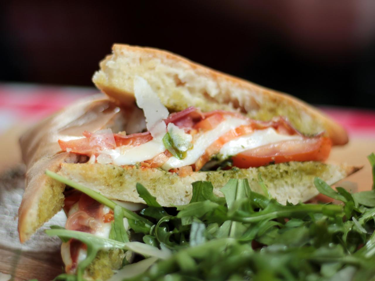 Kanapka z ciabatty z pesto, mozzarellą, pomidorem, szynką parmeńską i rukolą, posypana parmezanem, serwowana na drewnianej desce.