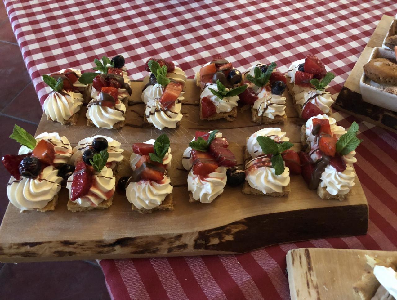 Rządki mini-deserów na drewnianej desce: kwadratowe ciasteczka z bitą śmietaną, truskawkami, borówkami, listkami mięty i polewą czekoladową, na tle czerwono-białego obrusu w kratę.