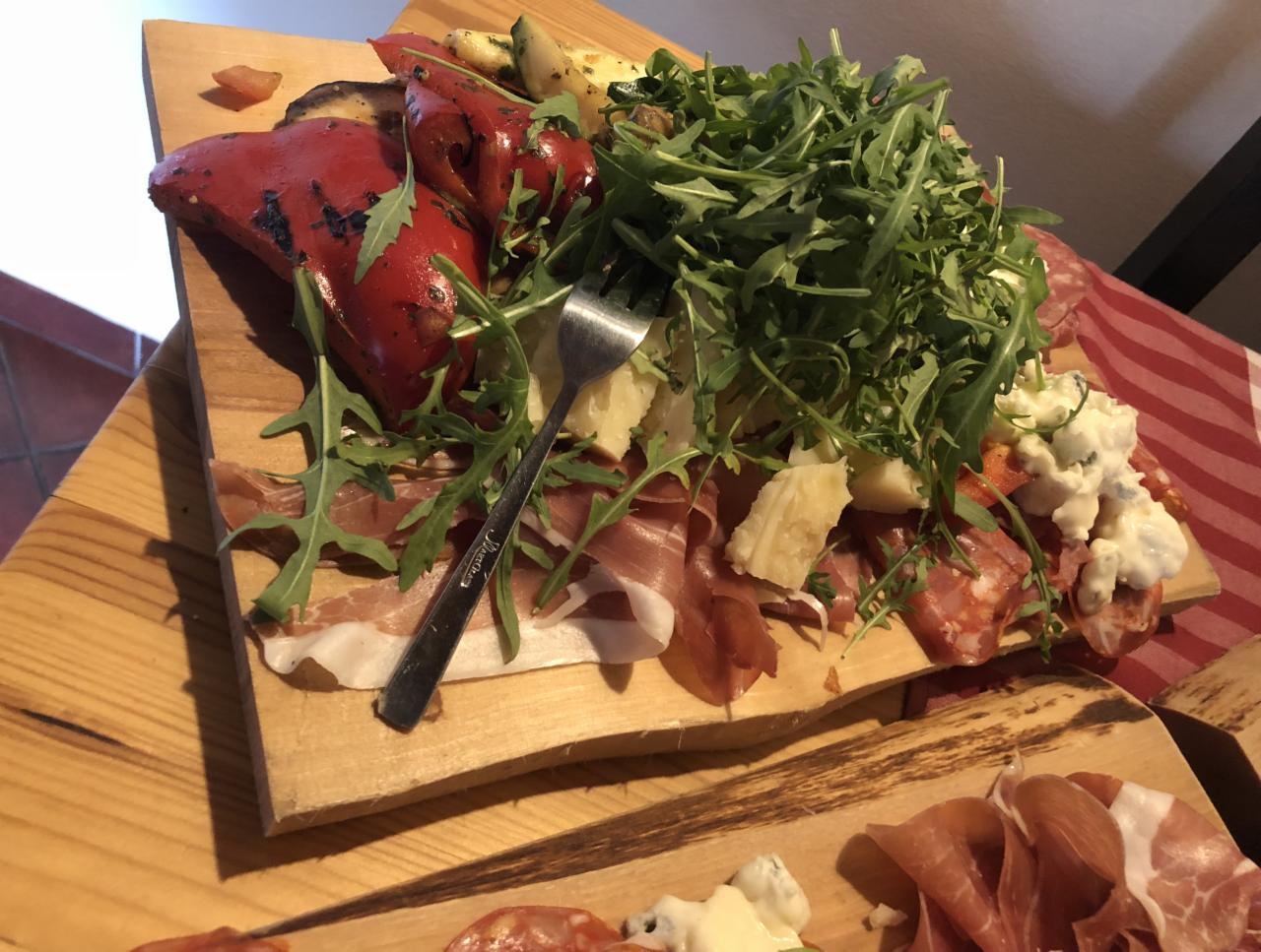 Drewniana deska z wędlinami, serami, pieczoną papryką, cukinią i rukolą, uzupełniona widelcem, na tle czerwono-białej serwety.