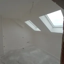 Poddasze po remoncie, świeżo wykończone ściany i sufit na biało, widoczne okna dachowe i fragment muru z cegły, na podłodze ślady po pracach wykończeniowych, elektryk w tle.