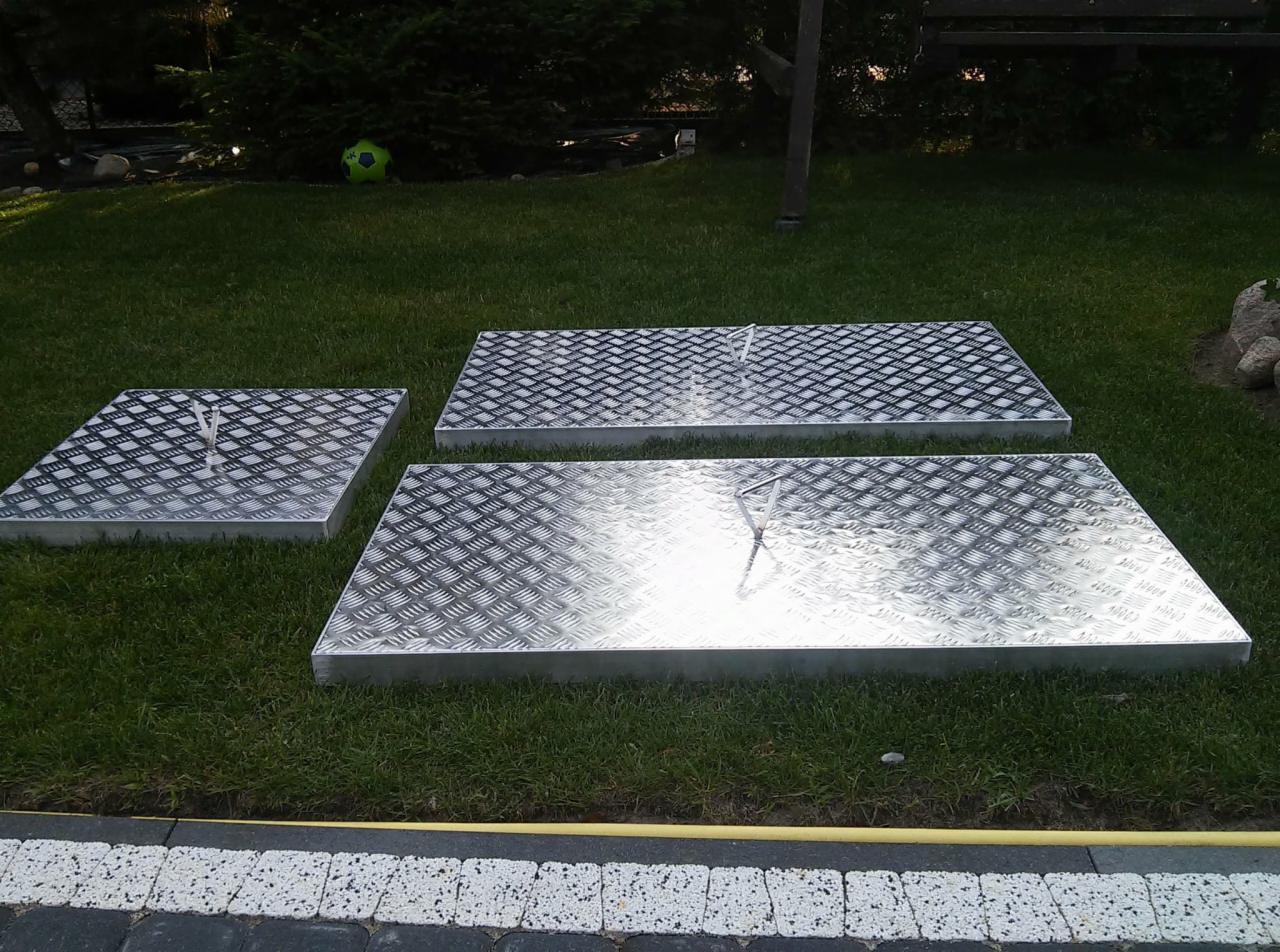 Trzy aluminiowe, antypoślizgowe pokrywy włazów z uchwytami, leżące na trawie w ogrodzie.