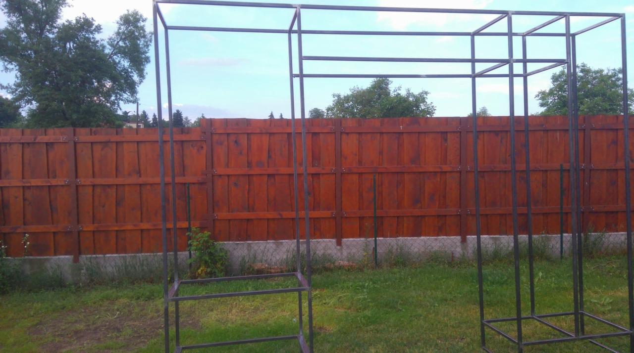 Metalowa konstrukcja szkieletowa, prawdopodobnie altany, na tle drewnianego ogrodzenia i zielonego trawnika.