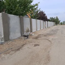 Świeżo wylany, surowy betonowy płot modułowy z widocznymi śladami szalunku, taczka z narzędziami i zaprawą obok, ziemista droga w tle.