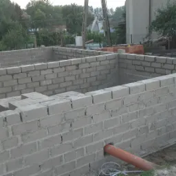 Wznoszenie ścian fundamentowych z bloczków betonowych, widoczny pomarańczowy rurociąg w fundamencie, teren budowy w tle z zielenią i budynkami.