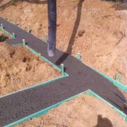 Wylewanie fundamentów pod budynek, widoczne szalunki z zielonych desek i rura do podawania betonu, teren budowy z ziemią.