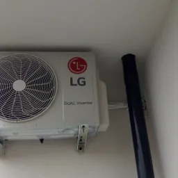 Zewnętrzna jednostka klimatyzacji LG Dual Inverter zamontowana na białej ścianie, widoczne przewody i rura odprowadzająca skropliny.