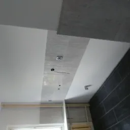 Wnętrze łazienki w trakcie prac wykończeniowych: częściowo ułożone płytki na ścianie i suficie, widoczne przewody elektryczne, ściany pomalowane na biało, ciemne płytki ułożone w cegiełkę.