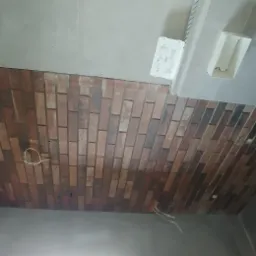 Ściana w łazience wyłożona płytkami imitującymi czerwoną cegłę, widoczne przewody elektryczne, fragment umywalki i pojemnik z zaprawą.