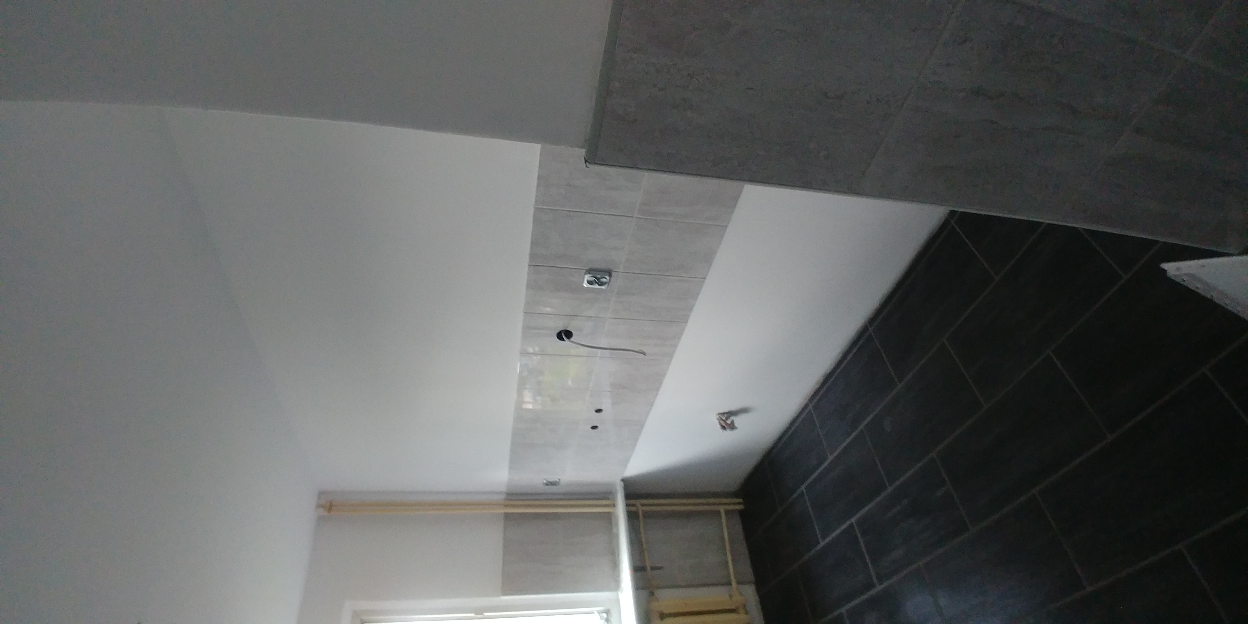 Wnętrze łazienki w trakcie prac wykończeniowych: częściowo ułożone płytki na ścianie i suficie, widoczne przewody elektryczne, ściany pomalowane na biało, ciemne płytki ułożone w cegiełkę.