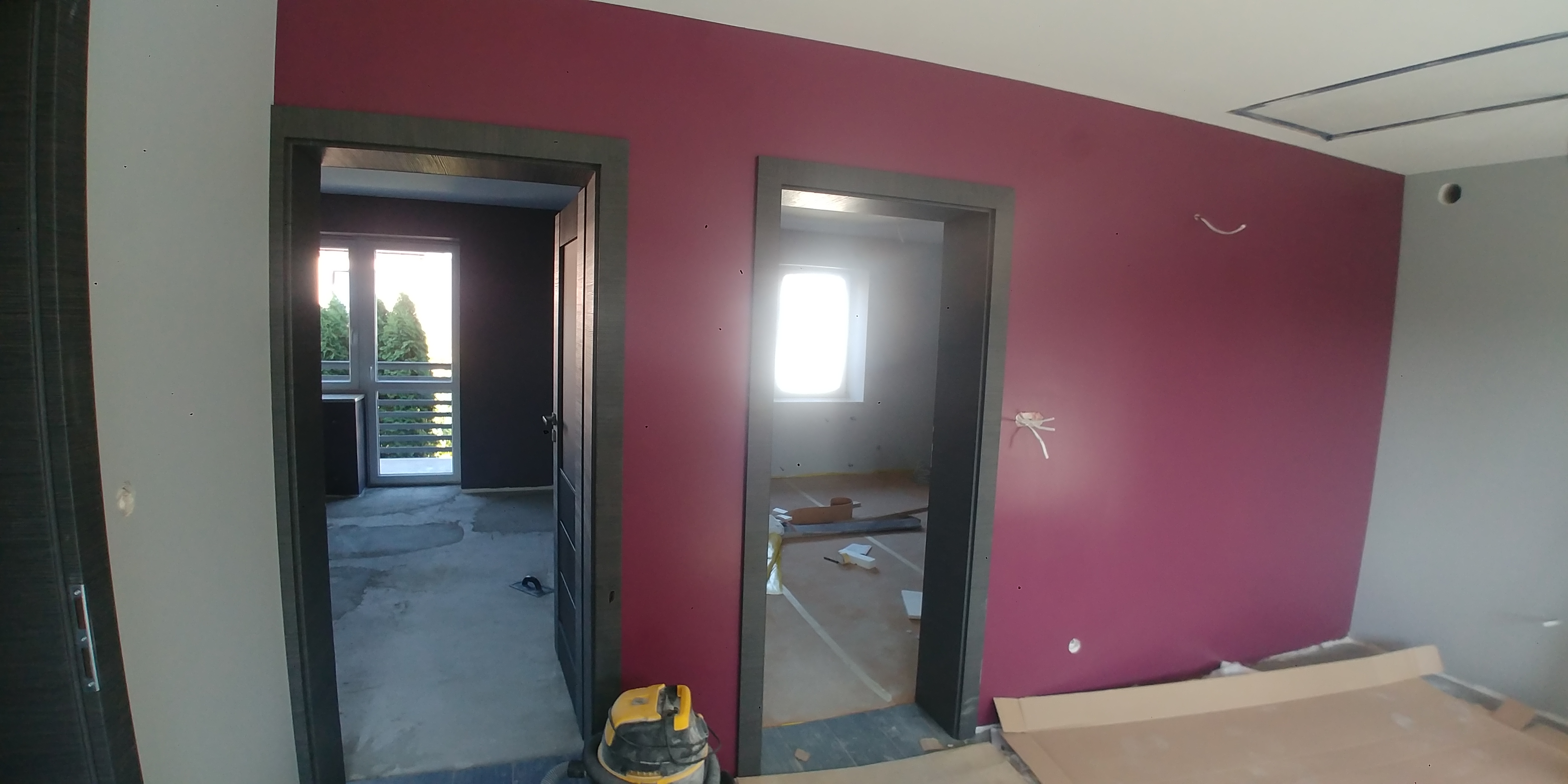 Wnętrze w trakcie remontu: pomalowana na burgundowo ściana, widoczne dwa otwory drzwiowe z szarymi ościeżnicami, podłoga zabezpieczona kartonem, odkurzacz budowlany w rogu.