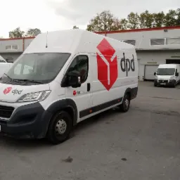 Biały samochód dostawczy DPD na tle magazynu z bramami, zaparkowany na betonowym placu w pochmurny dzień. Widoczny numer rejestracyjny RZ 0715T.