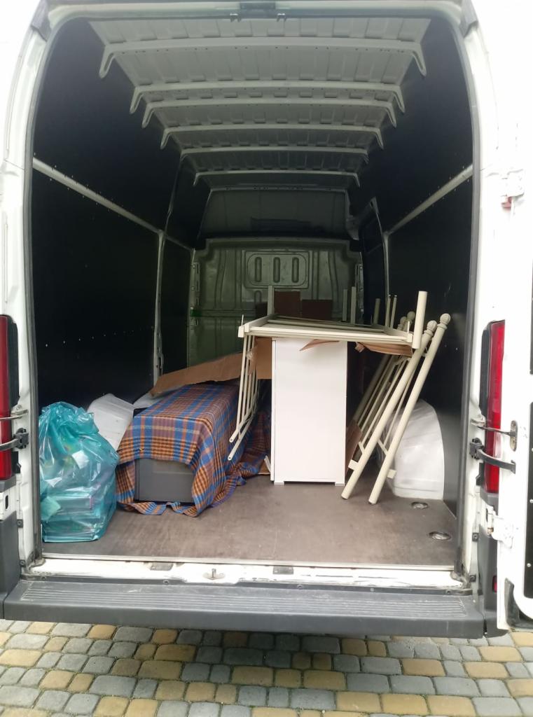 Wnętrze białego busa załadowane meblami i spakowanymi rzeczami, widoczne z tyłu pojazdu, na podłodze leżą przedmioty przykryte pledem w kratę, a po prawej stronie oparta jest rozłożona biała kołyska.