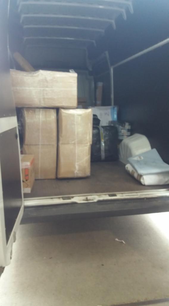 Wnętrze furgonetki wypełnionej kartonami i torbami, przygotowanymi do transportu. Widoczne zabezpieczenia na paczkach.