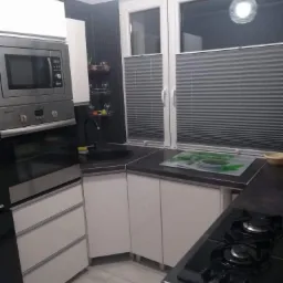 Nowoczesna kuchnia z białymi szafkami, czarnym blatem i okapem, z widocznymi plisowanymi roletami w oknie i czarną ścianą w tle.