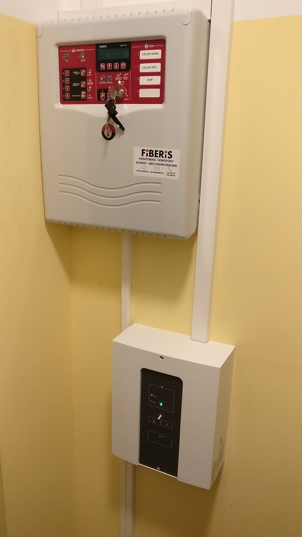 Centrala alarmowa z kluczykiem w stacyjce, zamontowana na żółtej ścianie, z widoczną dolną skrzynką sterującą i maskującymi listwami.