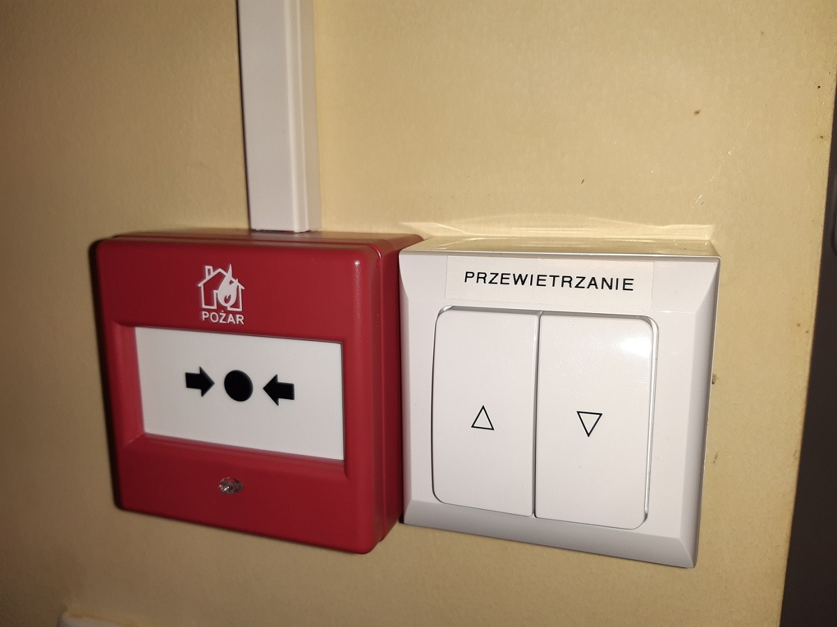 Czerwony przycisk alarmowy POŻAR z symbolem płomienia obok białego przełącznika PRZEWIETRZANIE z symbolami strzałek góra/dół na beżowej ścianie.
