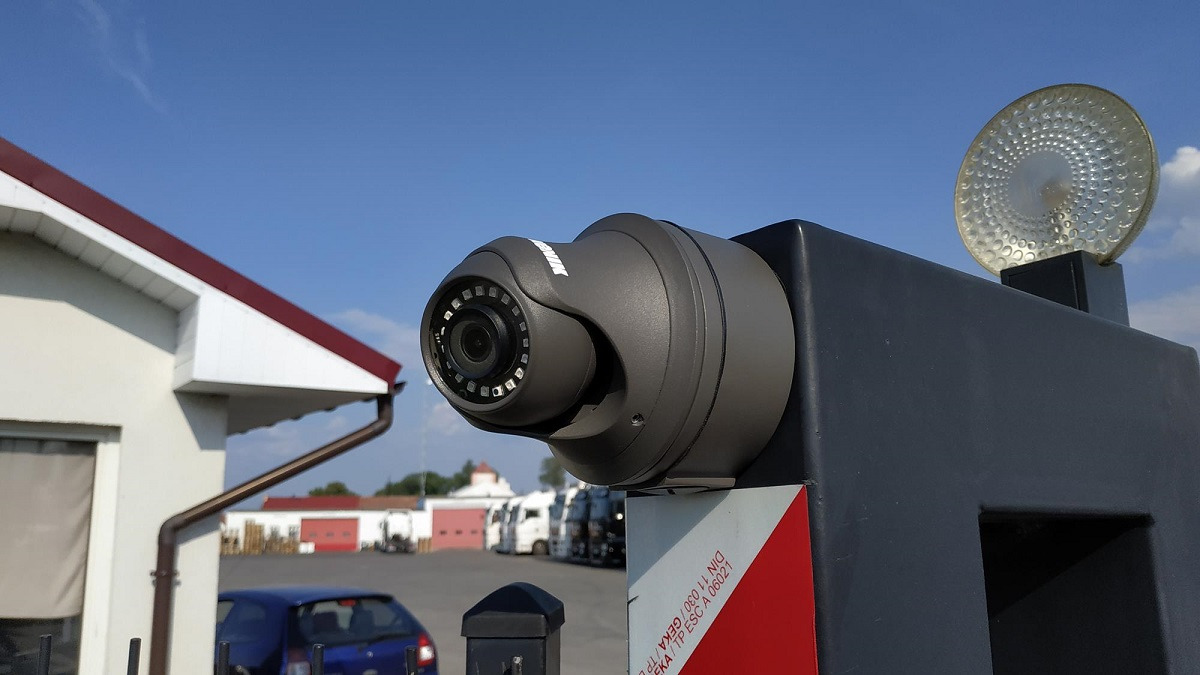 Kopułkowa kamera monitoringu zewnętrznego z widocznym obiektywem i diodami IR, zamontowana na czarnym słupku, w tle budynki i zaparkowane pojazdy w słoneczny dzień.