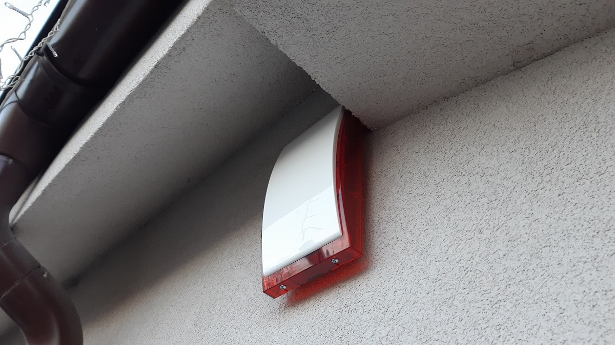 Zewnętrzna syrena alarmowa z czerwoną obudową zamontowana pod okapem budynku o szarej, chropowatej elewacji.