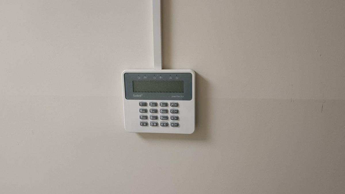 Biały panel sterowania alarmem Safel Perfecta zamontowany na jasnej ścianie, z widoczną listwą kablową u góry.
