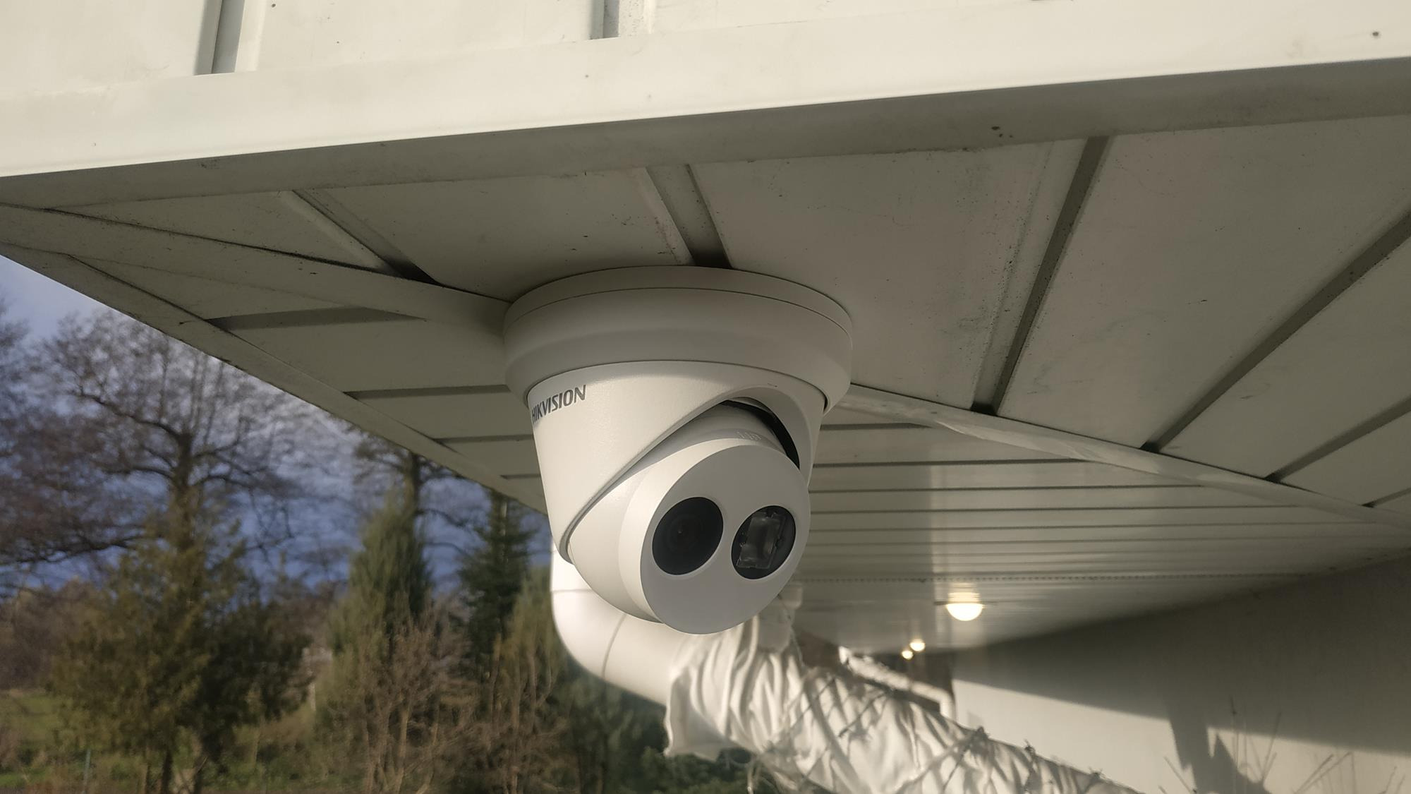 Kamera kopułkowa marki Hikvision zamontowana pod okapem budynku, widok z dołu na tle drzew i nieba.