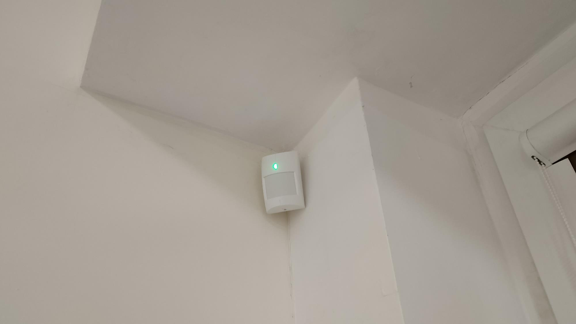 Biały czujnik ruchu z zieloną diodą LED zamontowany w rogu pomieszczenia na białej ścianie, widoczny fragment rolety okiennej.
