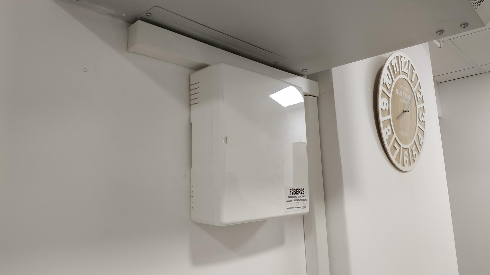 Biała centrala alarmowa Fiberis zamontowana na ścianie w pomieszczeniu biurowym, widoczny zegar w tle.