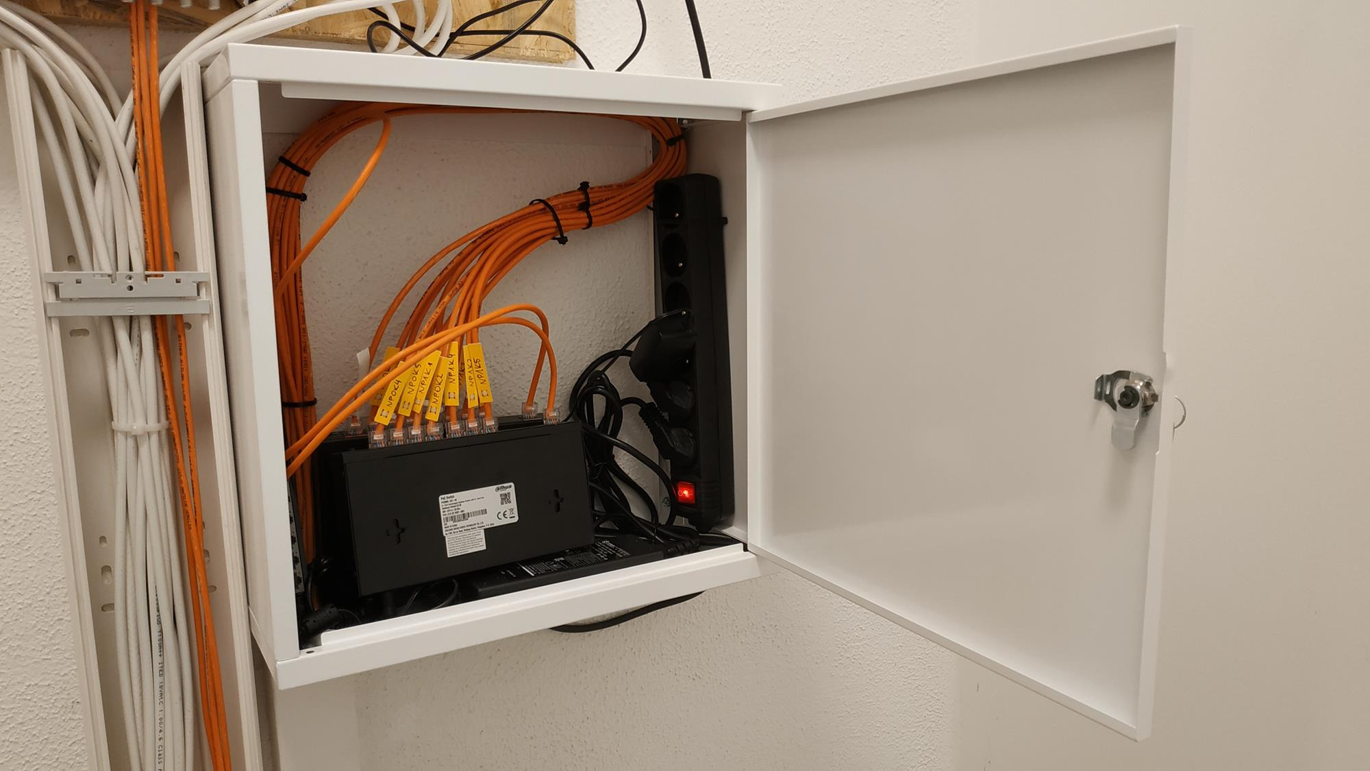 Otwarta biała szafka telekomunikacyjna na ścianie, widoczne pomarańczowe i białe kable, patch panel z oznaczonymi portami, listwa zasilająca z włączonym czerwonym wskaźnikiem.