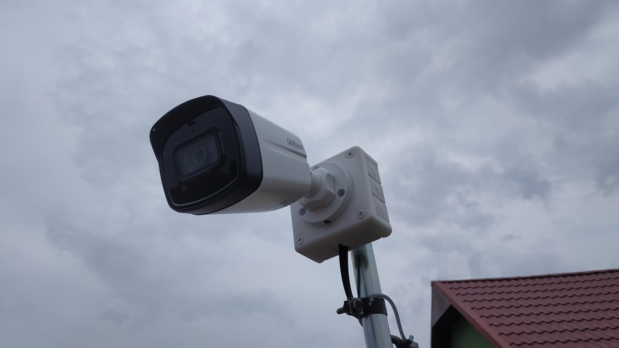 Kamera monitoringu Dahua zamontowana na metalowym słupku na tle pochmurnego nieba i fragmentu dachu z czerwonej dachówki.