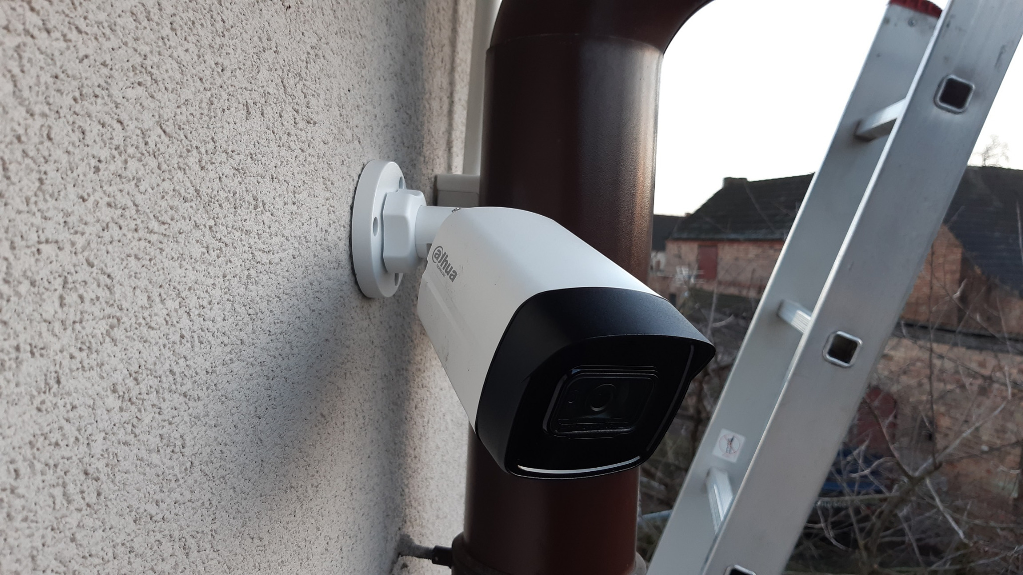 Biała kamera Dahua zamontowana na szarej, chropowatej ścianie budynku, obok brązowej rury spustowej i drabiny aluminiowej.