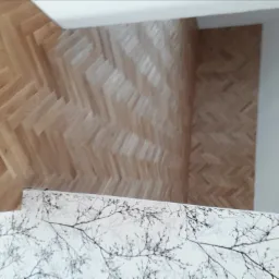 Ukośne ujęcie parkietu ułożonego w jodełkę z jasnego drewna, częściowo przykrytego białym blatem z wzorem gałązek.