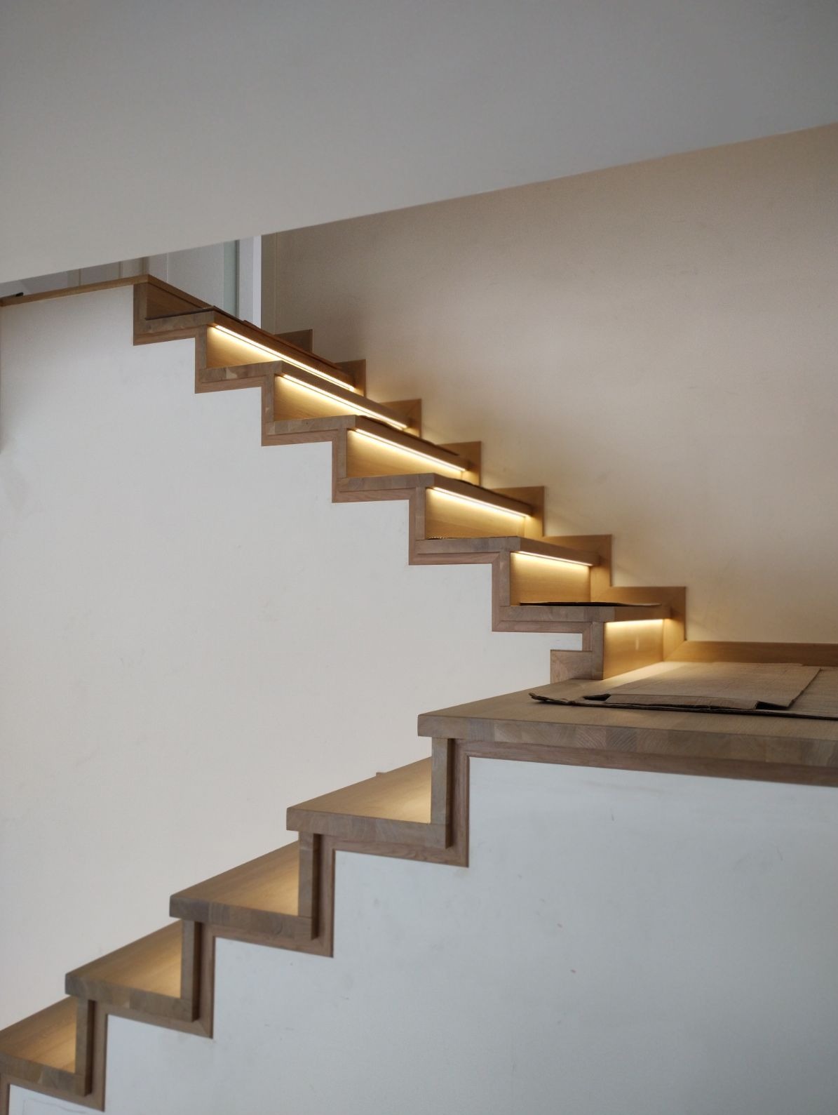 Drewniane schody z oświetleniem LED zamontowanym pod każdym stopniem, widoczne białe ściany i fragment sufitu, na jednym ze stopni leżą kartonowe arkusze ochronne.