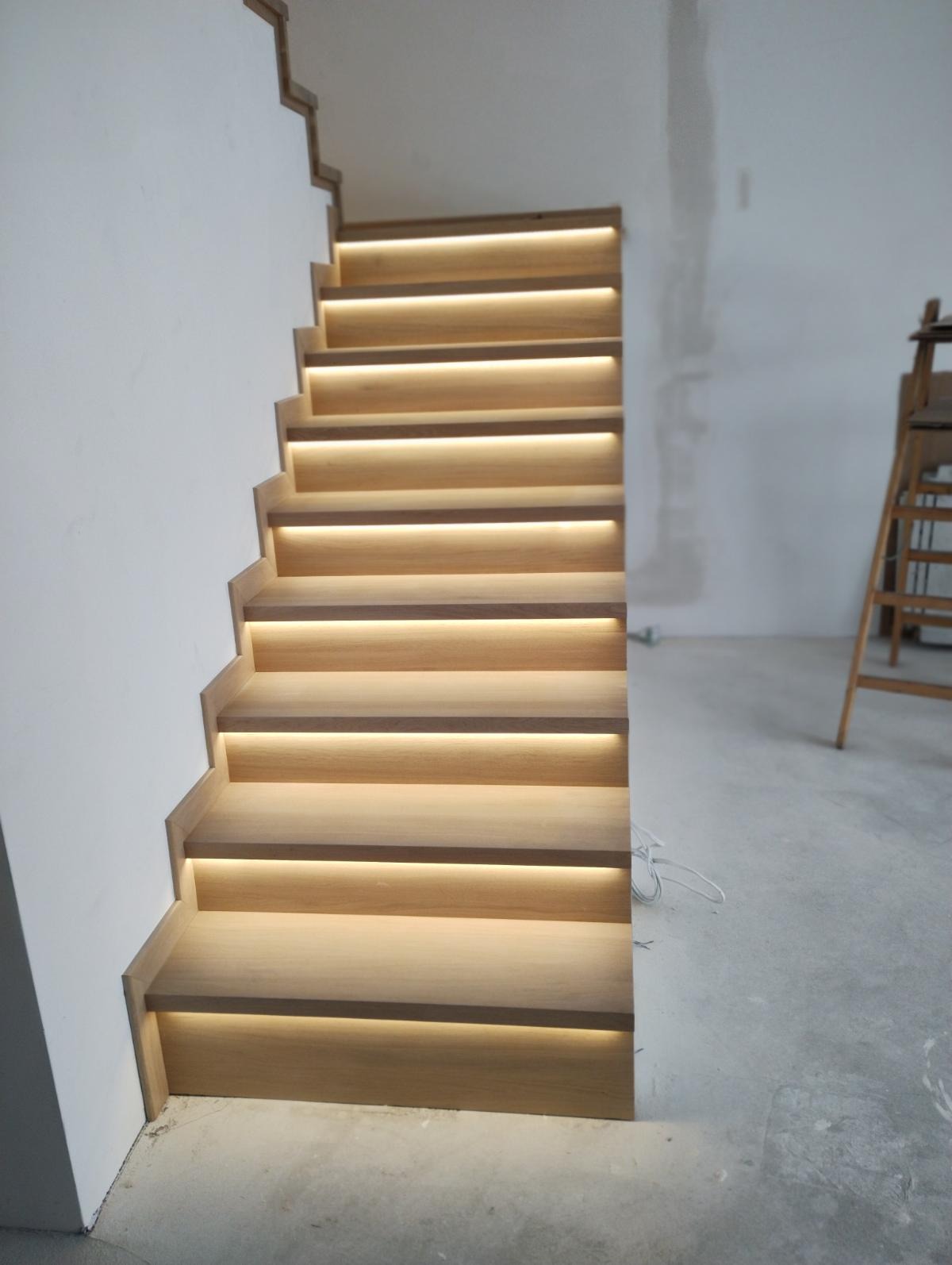 Drewniane schody z podświetleniem LED na każdym stopniu, widoczne kable elektryczne na podłodze, drabina w tle, surowy stan wykończenia ścian.