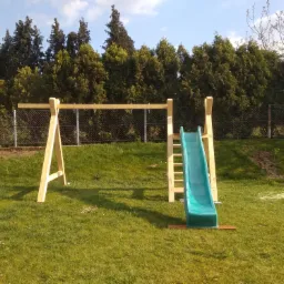 Drewniana konstrukcja huśtawki i zjeżdżalni na trawniku przed domem, widoczne drzewa w tle, częściowo zmontowana konstrukcja, świeże ślady montażu w trawie.