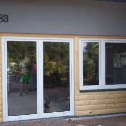 Nowe białe okna z widocznym odbiciem osoby i psa, zamontowane w drewnianej elewacji z numerem domu 83.