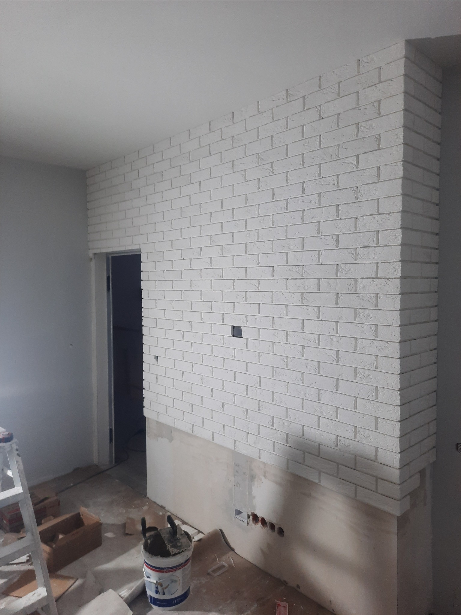 Ściana wykończona białą cegłą dekoracyjną, częściowo położona, widoczne puszki elektryczne, drabina i wiadro z zaprawą w pomieszczeniu w trakcie remontu.