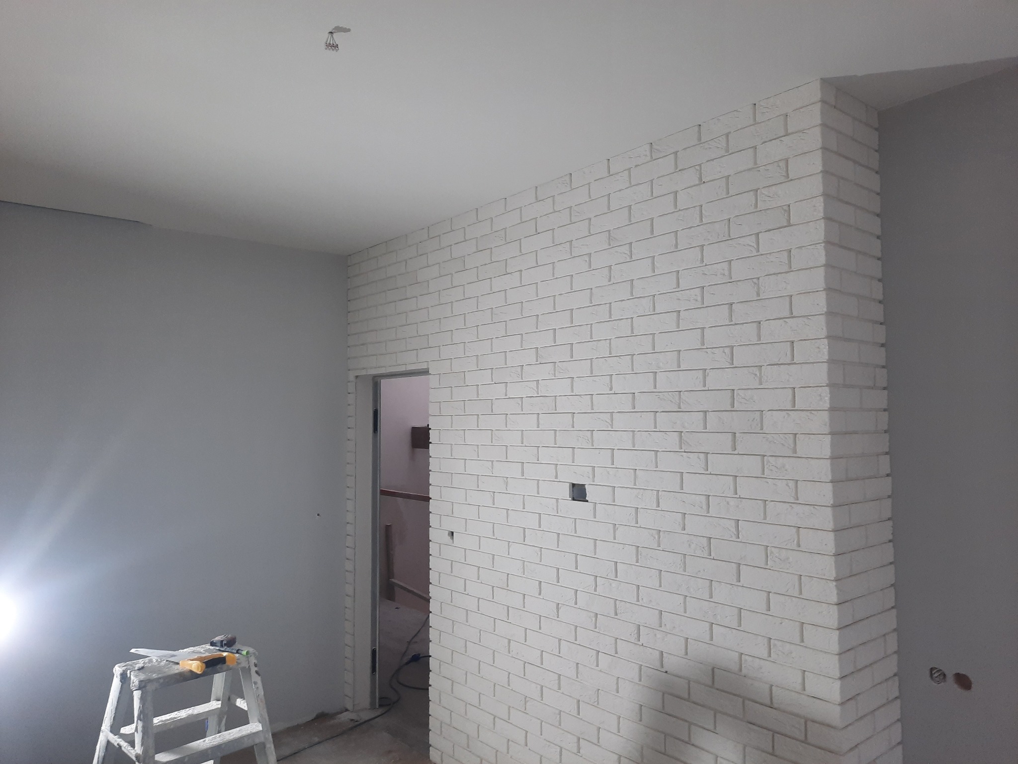 Ściana wykończona białą cegłą dekoracyjną, widoczna drabina malarska z narzędziami, szara ściana w tle, surowy stan wykończenia wnętrza.