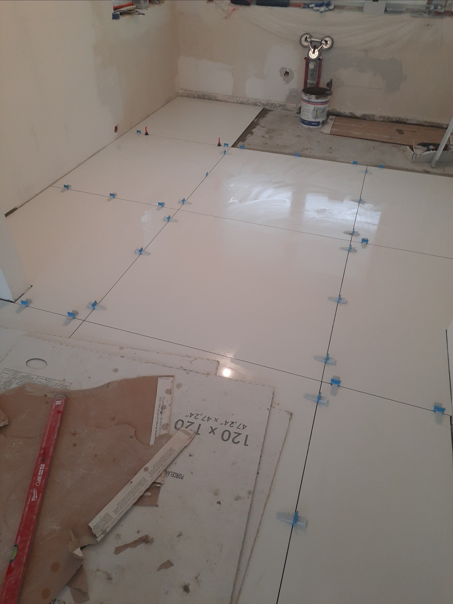 Układanie jasnych, wielkoformatowych płytek podłogowych z użyciem poziomicy, krzyżyków dystansowych i przyssawki do przenoszenia. Częściowo wykończona podłoga w pomieszczeniu w trakcie prac...