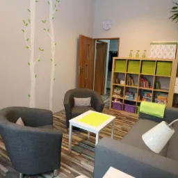 Gabinet z dwoma szarymi fotelami, sofą, białym stolikiem z żółtym blatem, regałem z książkami i zabawkami, dekoracją ścienną z motywem drzew i zegarem.