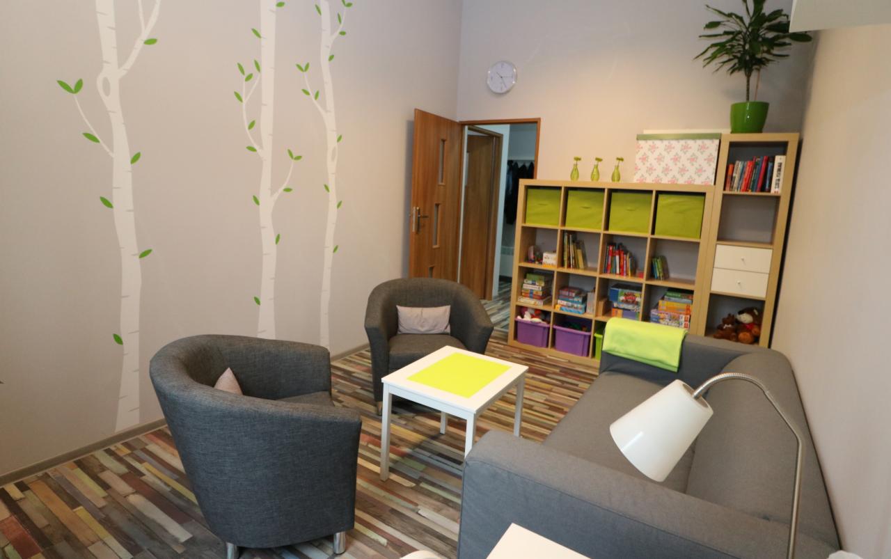 Gabinet z dwoma szarymi fotelami, sofą, białym stolikiem z żółtym blatem, regałem z książkami i zabawkami, dekoracją ścienną z motywem drzew i zegarem.