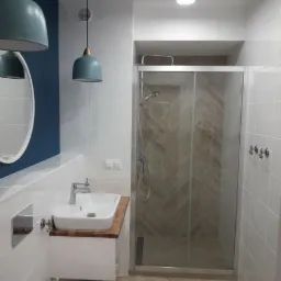 Nowoczesna łazienka z kabiną prysznicową ze szklanymi drzwiami przesuwnymi, umywalką nablatową na drewnianym blacie, wiszącą toaletą i dwoma turkusowymi lampami wiszącymi.