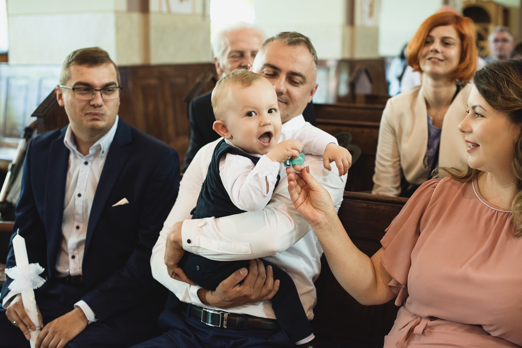Uśmiechnięty niemowlak w czarnej kamizelce trzymany na rękach przez mężczyznę, kobieta podaje smoczek, w tle goście w kościele.